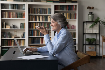 Older generation using modern tech, comfort communication, ai, workflow concept. Mature 60s business woman sit at workplace desk with laptop holds cellphone makes call talks to client on speakerphone