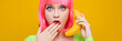 Young freaky surprised woman in a pink wig uses a banana as a phone on a bright yellow background