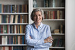 © fizkes - Happy middle-aged woman standing with arms crossed on bookshelves background, staring at camera look confident feels satisfied. Portrait teacher, library worker, professional occupation person concept
