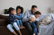 © fizkes - Positive cheerful Black mom and dad cuddling, tickling two children on sofa giggling, laughing, enjoying family fun, home activities, leisure, being parents. African couple playing with preschool kids