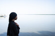 Lonely, troubled young African girl, dressed all in a black shawl and long robe, facing the sea, hands crossed on her chest in an anxious gesture; Loneliness and depression symbol