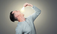 A Man Tries To Put A Lit Electric Light Bulb In His Mouth On Grey Background. 