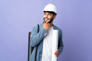 Young architect Moroccan man with helmet and holding blueprints over isolated background looking up while smiling