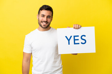 Wall Mural - Young handsome caucasian man isolated on yellow background holding a placard with text YES with happy expression