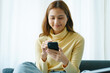 © Phushutter - Smiling young attractive Asian woman using her mobile phone while sitting on sofa at home.