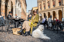 Stylish Woman Sitting With Her White Dog At Outdoor Cafe In The Old Town Of Bologna City. Italian Measured Lifestyle And Street Fashion Concept. Idea Of Traveling Italy
