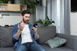 © Liubomir - Thinking man looking at phone screen sitting on sofa at home
