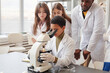 © Seventyfour - Portrait of young black girl looking into microscope with while doing experiments with group of children in school chemistry lab