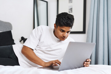 Wall Mural - Nothing passes the time like some cyber stalking. Cropped shot of an young man using his laptop while lying on his bed.