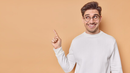 Happy positive adult man with dark hair smiles and points index finger above shows blank space to place your advertisement wears round spectacles casual jumper poses against brown background