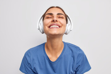 Wall Mural - Photo of pleased satisfied woman keeps eyes closed from pleasure catches every bit of song listens music via wireless headphones smiles toothily wears casual blue t shirt poses against white wall