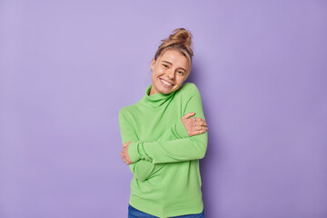 Sticker - Pleased lovely woman with combed hair embraces herself feels comfortable wears green jumper smiles gently being self egoist poses against purple background. Self love and acceptance concept.