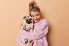 Display Of Affection. Happy Young Woman With Combed Hair Embraces Pug Dog Has Fun Being Mom Of Puppy Wears Knitted Sweater Isolated Over Beige Background. Female Pet Lover With Domestic Animal