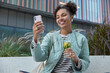 © wayhome.studio - Outdoor shot of happy teenage girl with two curly buns takes selfie via smartphone drinks detox beverage wears casual striped jumper and jacket carries net bag spends free time in urban setting