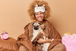 © WHstudio Leushin N - Positive smiling curly haired woman wrapped in blanket carries pug dog on hands stays in bed going to have sleep enjoys cozy domestic atmosphere poses against beige background. Family time concept
