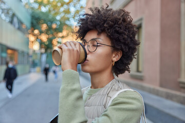 Wall Mural - Lovely curly haired young woman drinks takeaway coffee enjoys aromatic caffeine beverage wears spectacles casual clothes poses outdoors at street against blurred background. Lifestyle concept