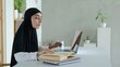 © Sia - A Muslim female teacher reads a book and types learning material on a laptop keyboard. The rights of Muslim women to education. A woman in a black hijab conducts remote training.