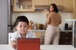 © DragonImages - Serious little boy watching educational video on tablet computer when his mother cooking dinner in background