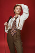 Fashionable beautiful singer girl in retro coat, tie and pants with microphone in hands posing on red background. Vintage toning.