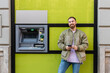 © Westend61 - Smiling young man with smart phone standing by atm