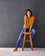 Positive And Carefree. Portrait Of A Happy Young Woman Sitting On A Stool Against A Gray Wall.