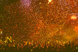 Fototapeta Dmuchawce - A crowded concert hall with scene stage orange and yellow lights, rock show performance, with people silhouette, colourful confetti explosion fired on dance floor air during a concert festival