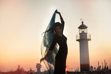 A Young Woman On The Background Of A Lighthouse During Sunset On The Seashore. Beautiful Slender Silhouette Of A Female Figure With Developing Hair.
