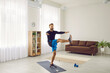 © Studio Romantic - Fit man having active workout at home. Handsome strong athlete following individual fitness program doing leg swing exercise on sports mat and using modern mobile phone app to track physical activity