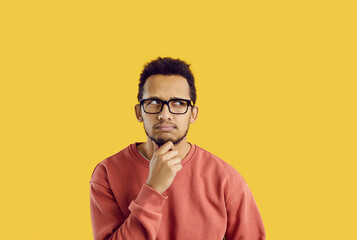Hmm. Confused young student thinking hard. Funny puzzled adult man in glasses and casual sweatshirt looking up on solid yellow background, rubbing his chin, thinking, planning, doubting something