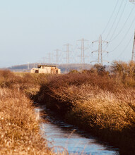 Small Creek Flowing Through A Grassland And A Building Near To Electric Power Poles Against Blue Sky