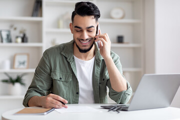 Wall Mural - Handsome Arab Male Freelancer Talking On Cellphone And Taking Notes At Home