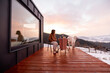 Woman with her dog resting on terrace of tiny house in the mountains, enjoying beautiful landscape during sunrise. Concept of small modern cabins for rest and escape to nature