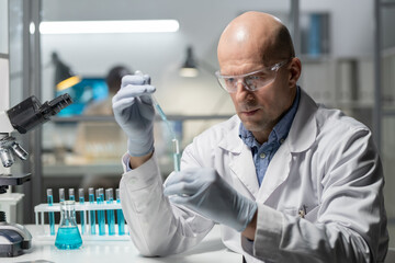 Poster - Mature experienced scientist in protective workwear mixing two chemical substances in flask while sitting by workplace in laboratory