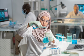 Aufkleber - Young serious Muslim female researcher in hijab, gloves and protective eyewear pouring liquid substance into flask with blue fluid