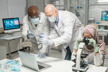 Aufkleber - Interracial male scientists discussing blue liquid in flask next to young Muslim female virologist studying new virus in microscope