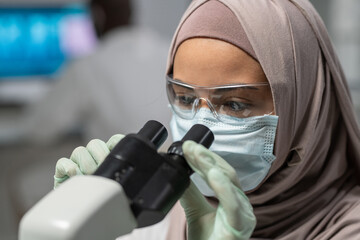 Fototapete - Close-up of young Muslim female researcher looking in microscope during scientific investigation or discovery of new virus