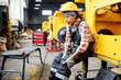 © pressmaster - Young female worker of warehouse in coveralls and hardhat touching hurting knee while sitting by industrial machine