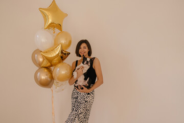 Wall Mural - Cheerful young european woman holding boston terrier looking at camera with smile on face, space for text. Dark-haired beauty stands near wall with balloons. Love between dog and owner concept