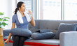 © Bangkok Click Studio - Asian young pretty happy healthy female athlete sport girl in casual sporty outfit sitting on sofa holding drinking bottle of water relaxing resting after learning exercising online in living room