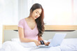 © Bangkok Click Studio - Asian young pretty female shopper in casual pajama waking up sitting smiling under blanket on bed in bedroom holding looking at credit card with light flare effect shopping online via laptop computer