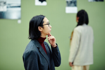 Young Asian man wearing eyeglasses standing in modern art gallery looking at photos at exhibition and thinking about something