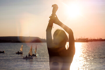 Silhouette of a strong confident woman on the seashore with a view of beautiful yachts. Portrait of a thin girl in a black dress with her hands raised at sunrise or sunset