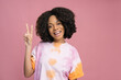 Young smiling Nigerian woman showing victory sign isolated on pink background, copy space. Happy curly haired female  wearing stylish tie dye t shirt posing for pictures in studio
