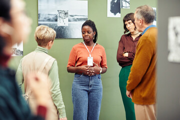 Portrait of young African American gallery worker standing in front of visitors discussing black and white photography