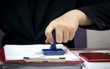 Man's Hand In A Stamp On A Document In A Company. Office Equipment, Approval, Registration Of Documents, Stamping For Symbols
