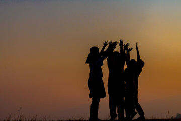 The silhouette of the children holding hands, enjoying the sunset, a group of friends cheering and arms raised in the orange sky and the mountains behind Friendship concept