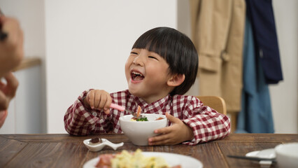 closeup of a cute Asian little kids boy holding spoon and bowl and looking at his family with a big smile laughing while having dinner together at dining table at home