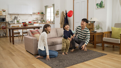 Wall Mural - excited asian baby boy playing with a red balloon with his father and mother in the living room at home. they hit the ball trying not to let it fall onto ground