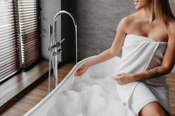 Wall Mural - Young lady wrapped in towel preparing to take bath, sitting on bathtub and touching water flowing from tap, crop