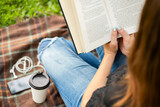 Fototapeta  - A woman in jeans with cup of coffee reading book while sitting on a blanket in the park
